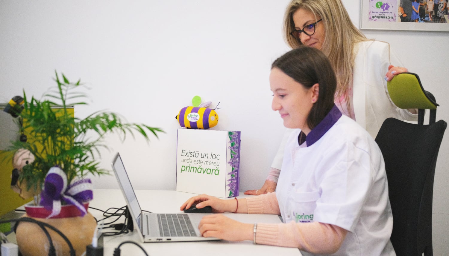 Farmacistă Spring FARMA în halat alb, alături de o colegă la laptop