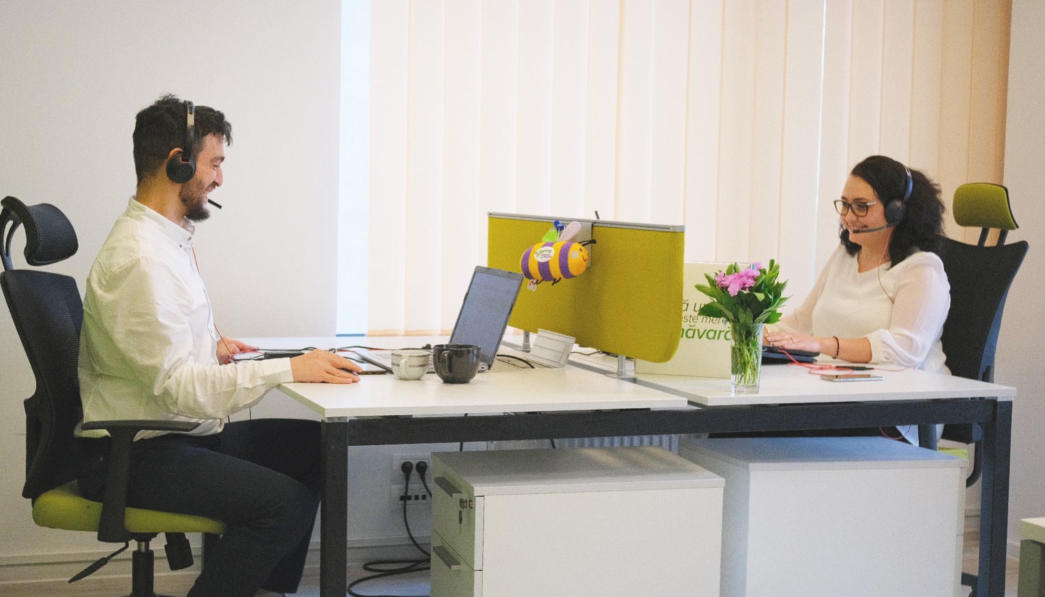 Doi agenți Customer Support cu căști audio, la birourile lor