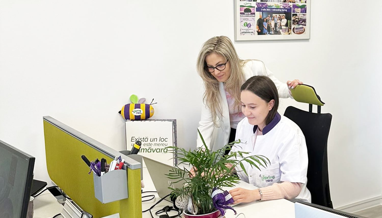 Farmacistă Spring FARMA în halat alb, ajutată de o colegă la laptop